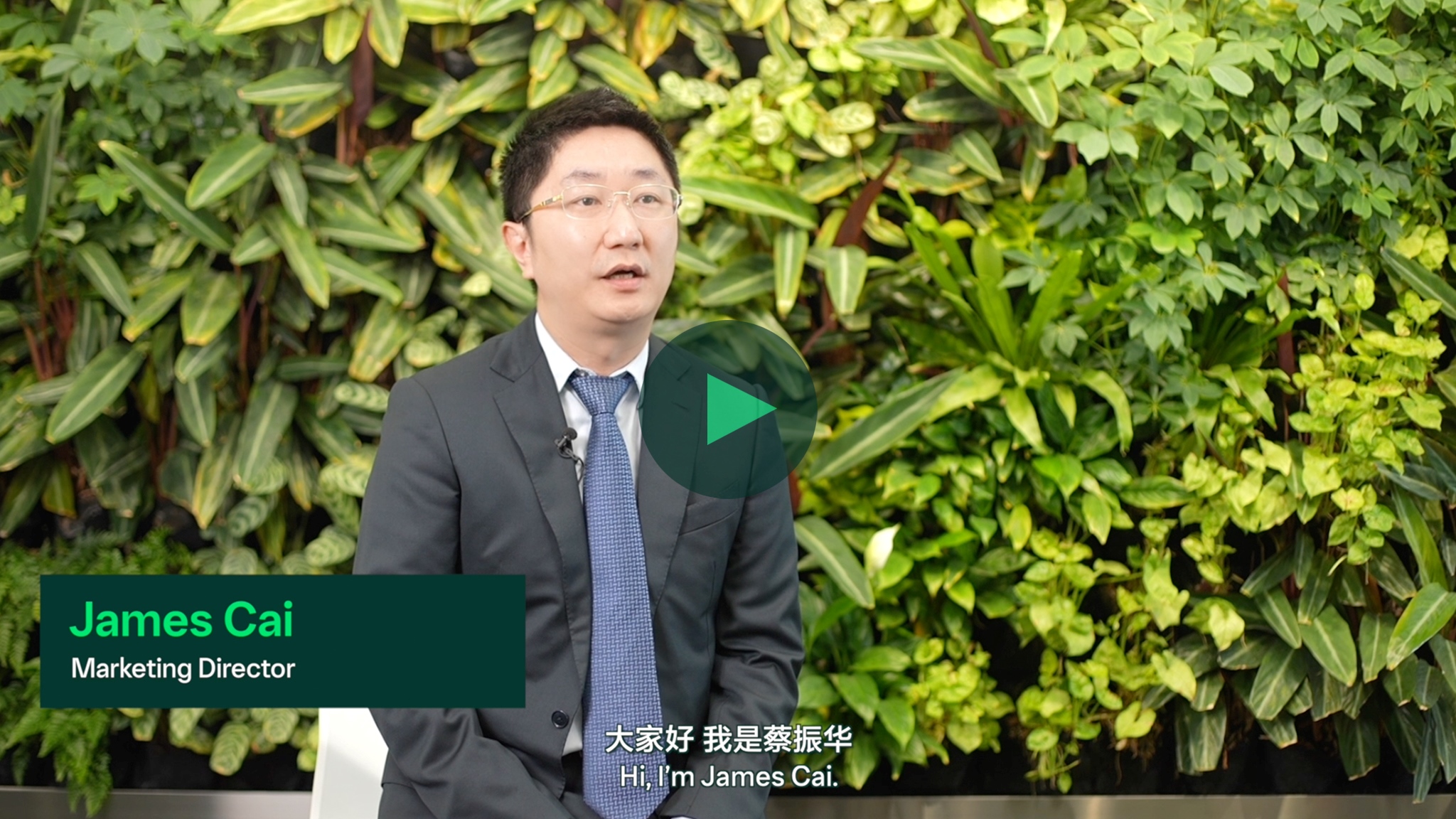 室内拍摄，一位身穿西装的人坐在绿色植物墙前，画面中有播放按钮。