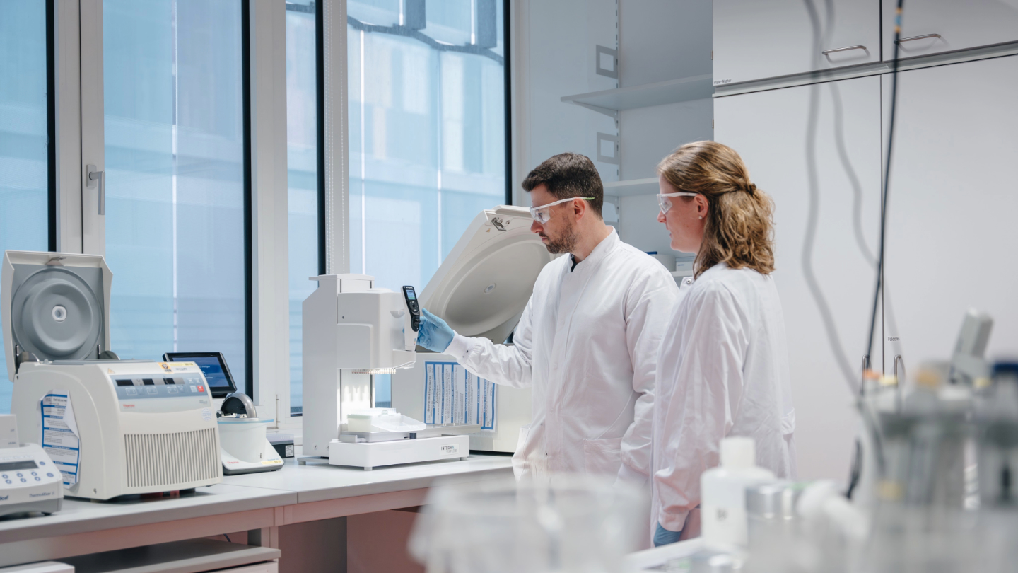 两位身穿白色实验服的科学家正在使用实验设备进行研究；镜头随后拉远，展现出实验室工作区域的整体环境。